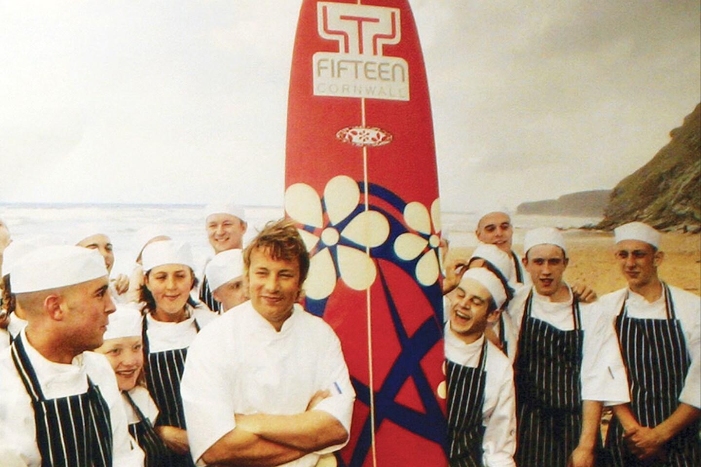 Jamie Oliver on the beach at Watergate Bay with a surfboard and apprentices