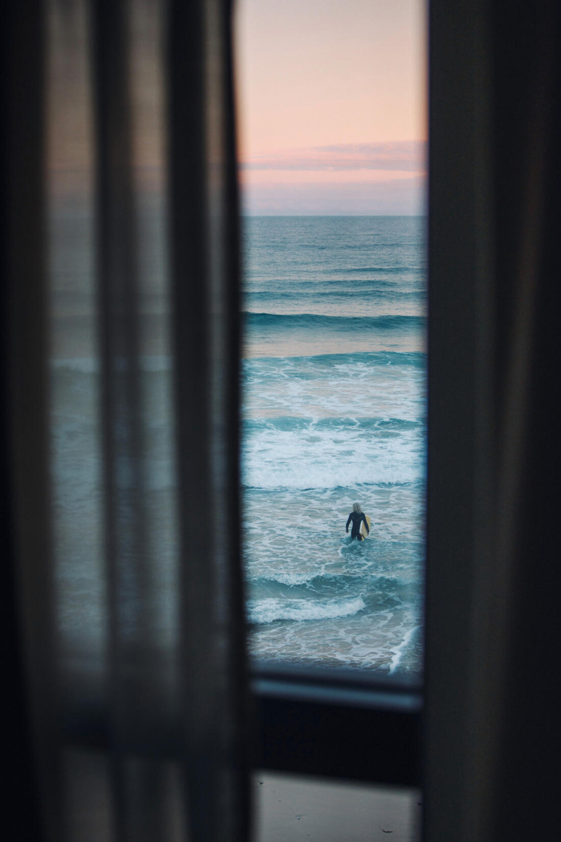 Curtains framing the photo with a view of a surfer walking into the sea with their surf board