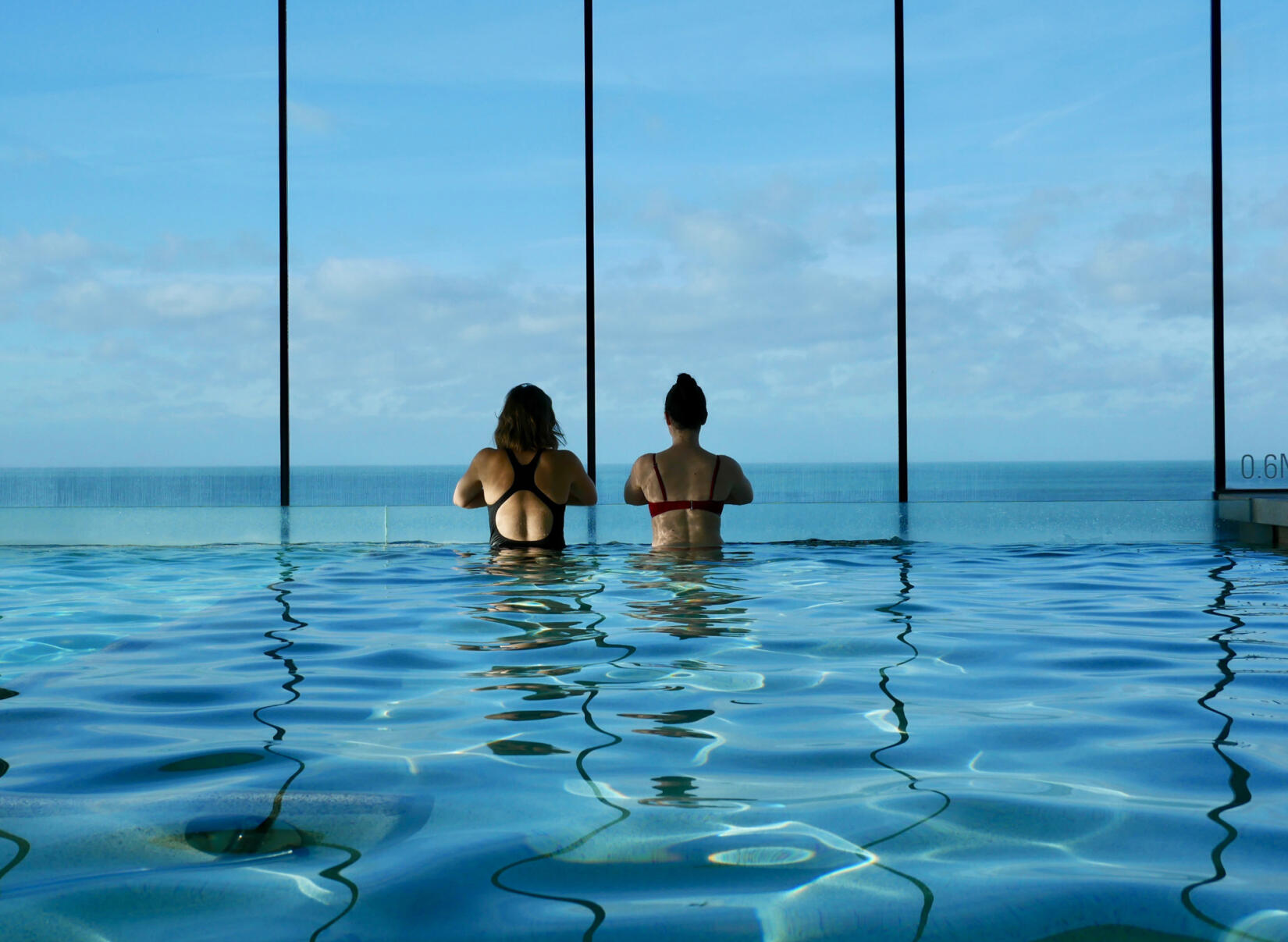 Two people stand looking out across the sea from the sea view pool in Swim Club at Watergate Bay