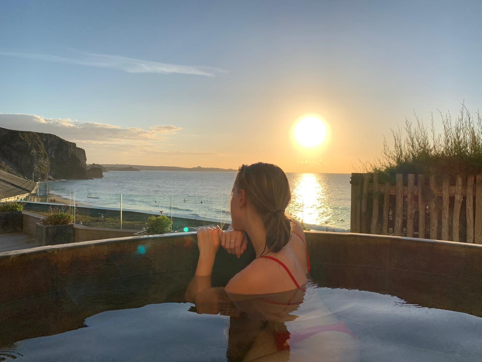 A lady in a hot tub facing towards the sea watching the sun set over the sea