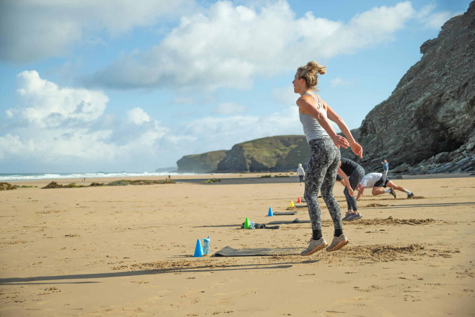 A HIIT class taking place on the beach with a lady jumping in the foreground