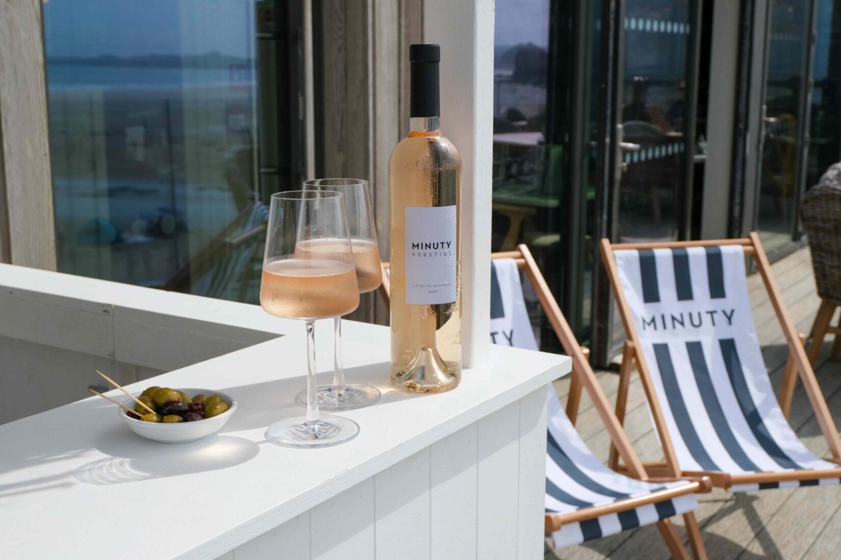 A bottle of minuty rose alongside two crisp glasses and a bowl of olives, minuty branded deckchairs in the background